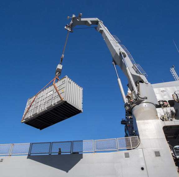 The crane on HMNZS Canterbury lifts a container onto the ship under blue skies.