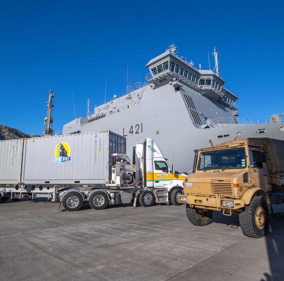 HMNZS Canterbury under blue skies with a commercial truck with grey containers on it (left) and a NZ Army Unimog (right)