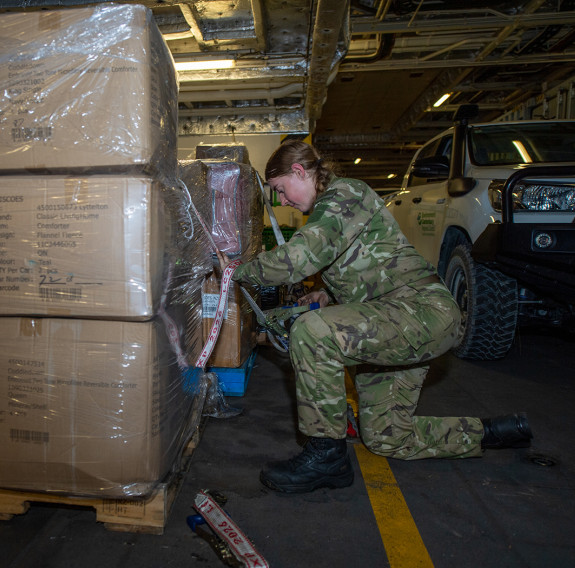A soldier in full camouflage and black boots pushing a pallet of cardboard boxes within HMNZS Canterbury.