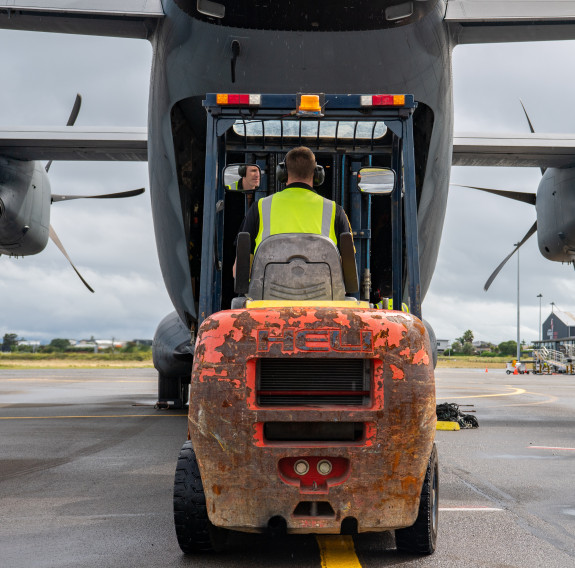 An aviator loads goods into the back of the RAAF C-27 Spartan using a forklift. On the right are reflective glass-fronted terminal buildings.