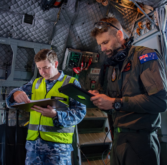 Two RAAF crew check a clipboard and iPad in the interior of the C-27 Spartan prior to their departure. The man on the left is wearing camouflage uniform and a high vis vest and the man on the right is wearing brown overalls with an Australian flag on his 