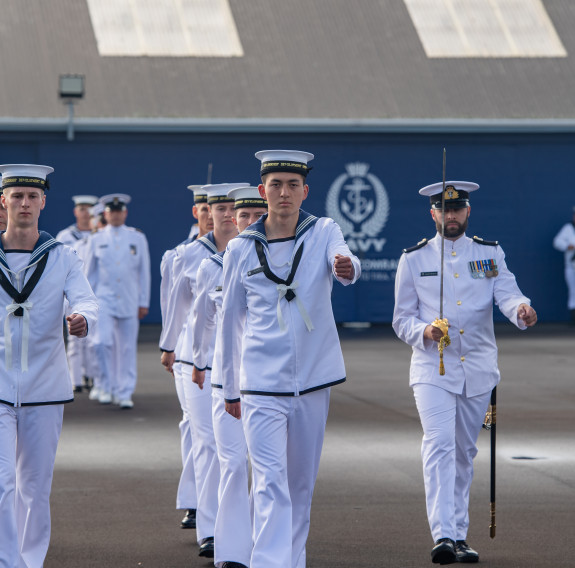 Sailors march on the parade ground in front of a blue building which has the RNZN emblem painted on the side. 
