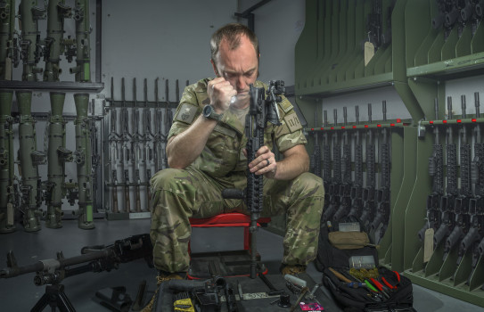 CPL Peden uses a torch to inspect a rifle in a room lined with green metal shelves, where rifles and accessories are stored.