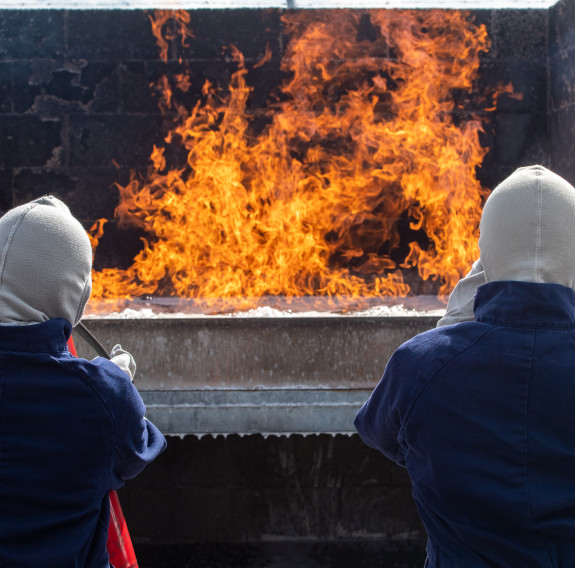 Two students confront controlled flames while wearing blue protective coveralls and face protection, equiped with fire extinguishers.