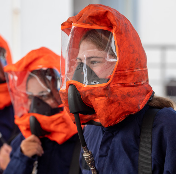Three students wear orange head coverings and respirator with clear visors while watching demonstrations.