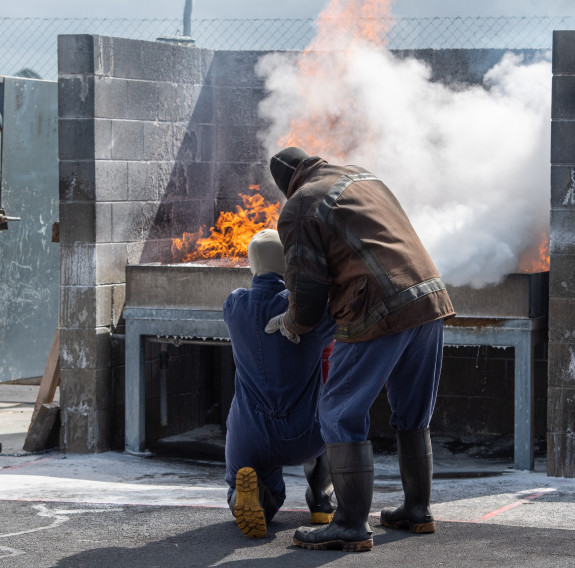 A student is instructed on how to extinguish a fire using fire extinguishers while wearing protective clothing.