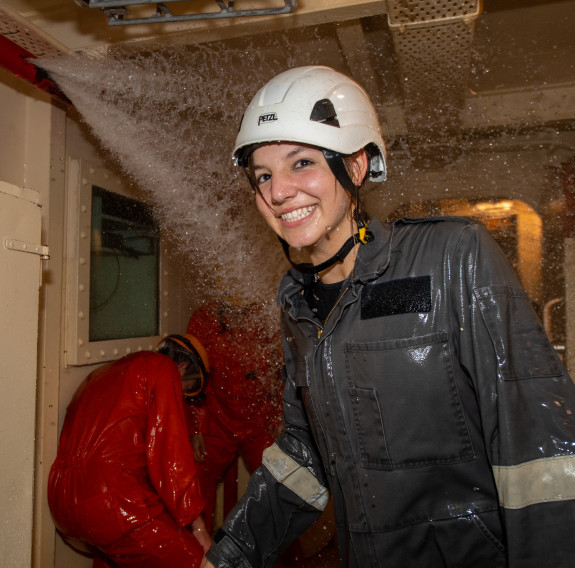 A student smiles at the camera after walking through sprays of water while wearing a while helmet and grey overalls.
