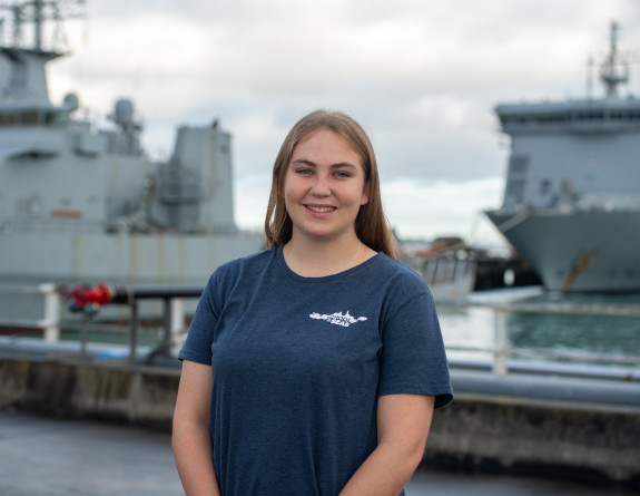 Shirleane Leuty, a School to Seas participant stands in front of two RNZN ships at Devonport Naval Base, she is wearing a blue 'School to Seas' branded t-shirt.