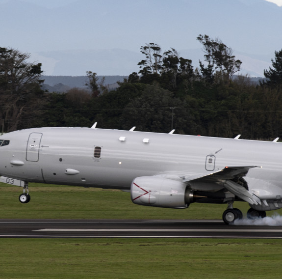 The grey P-8A touches down at Base Ohakea with smoke coming from the rear tyres as they make contact with the runway. The runway has grass on either side with trees in the background.