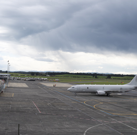 The P8-A taxis to in front of the hangers (left) while dark clouds form in the background. The apron is parked with white and yellow lines to indicate the path the aircraft should take.