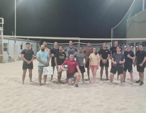 CPL Norman takes a knee in front of a large group of personnel on a sandy volleyball court on base in the Middle East.