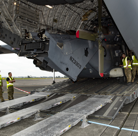 An NH-90 helicopter is loaded into the RAAF C-17, during the daytime. The helicopter is being loaded via the rear door and winched into position before it'll be secured for flight. Personnel in high visibility vests are in the aircraft and on the tarmac b