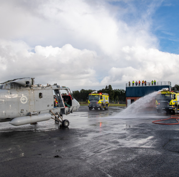 Two RNZAF Rescue vehicles at the scene of the simulated crash while personnel look on from an elevated viewing platform in the background.