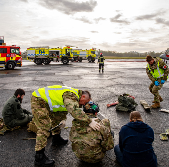 First responders treat the 'wounded' on the taxi-way of Base Auckland. FENZ and RNZAF vehicles in the background. Three people are sitting on the ground and two personnel tend to them.