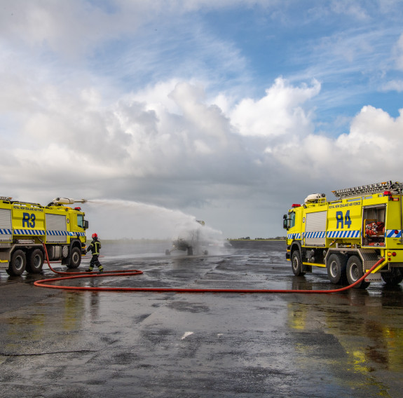 Each side of the frame is a RNZAF Fire Truck, in the middle of the frame, in the distance is the aircraft. The vehicle on the left sprays a large jet of water towards the simulated crash.