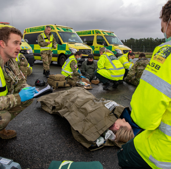 RNZAF personnel work with medics from Hato Hone St John to treat patients. They are positioned in front of three ambulances on an overcast day.