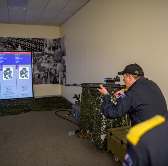 A sailor wearing a baseball cap and Navy uniform points the MARS-L weapon at a screen in an indoor environment.