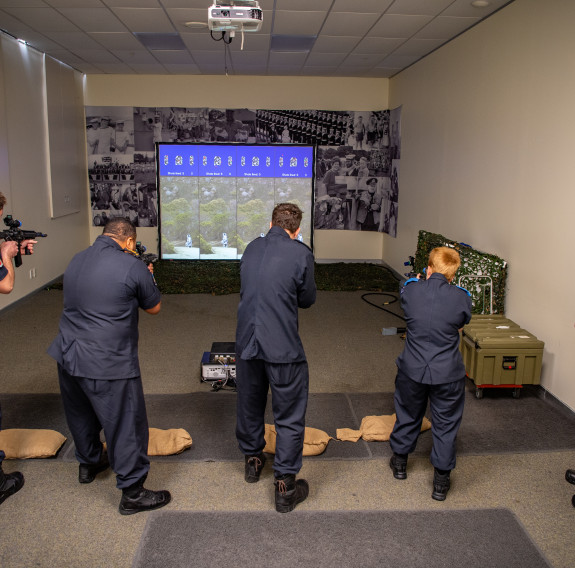 Four sailors have their back to the camera as they point their weapons down range at a screen. An instructor stands off the the right, observing.