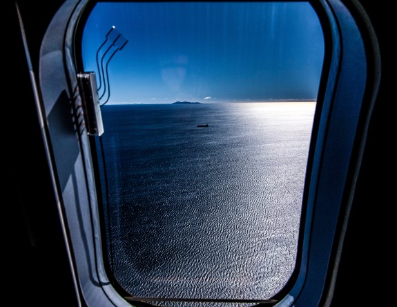 The view outside the window of a Royal New Zealand Air Force P-8A Poseidon aircraft that's flying over the water. In the ocean, you can see a ship and as well as an island.  