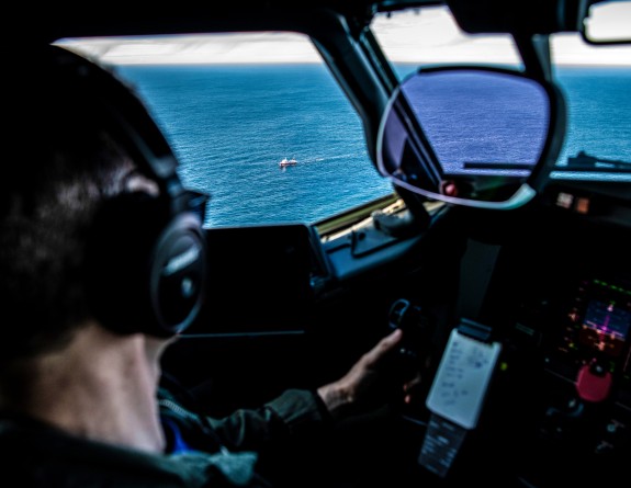 Photo taken over the shoulder of a Royal New Zealand Air Force pilot flying a P-8A Poseidon aircraft. The pilot is looking out the window at a ship in the ocean. 