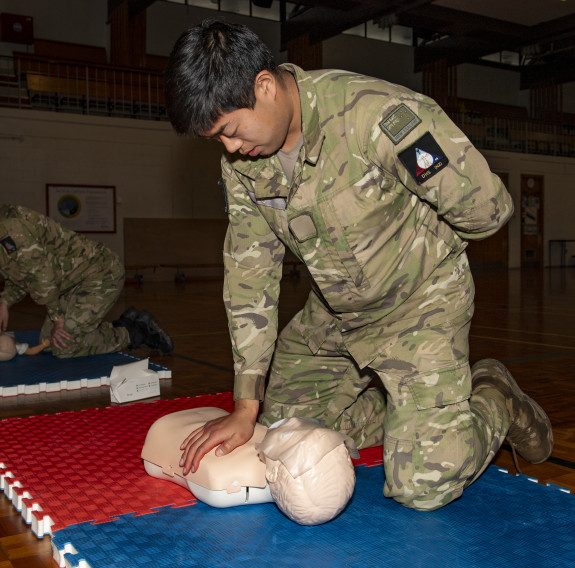A uniformed personnel performs CPR on a dummy. Another is in the background of the image. 