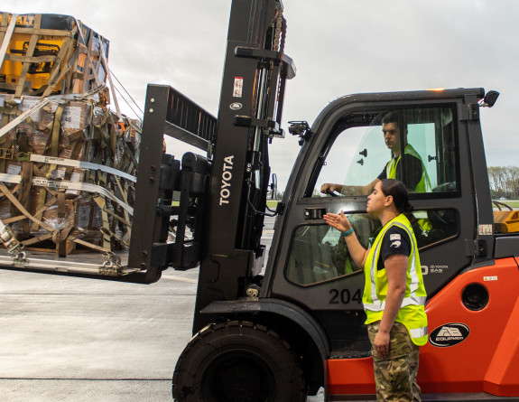 An person in camouflage Air Force uniform and a high visibility vest directs a forklift operator to move a pallet of supplies destined for Fiji.