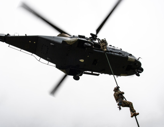 A military person winches down from an NH90 helicopter - the photo is taken looking up towards the helicopter.