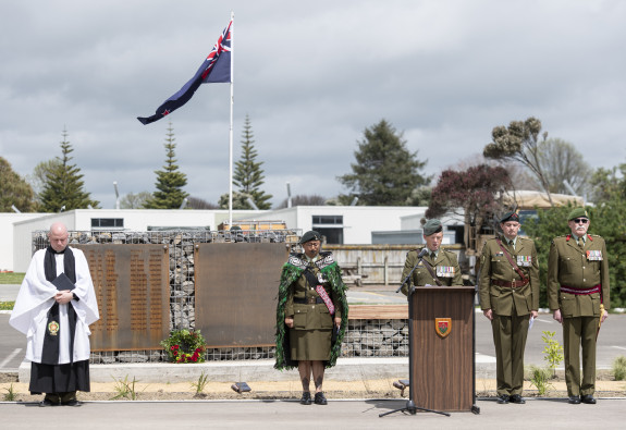 Five Army personnel stand in front of the memorial wall during the opening ceremony. The New Zealand flag is flying near the wall. 
