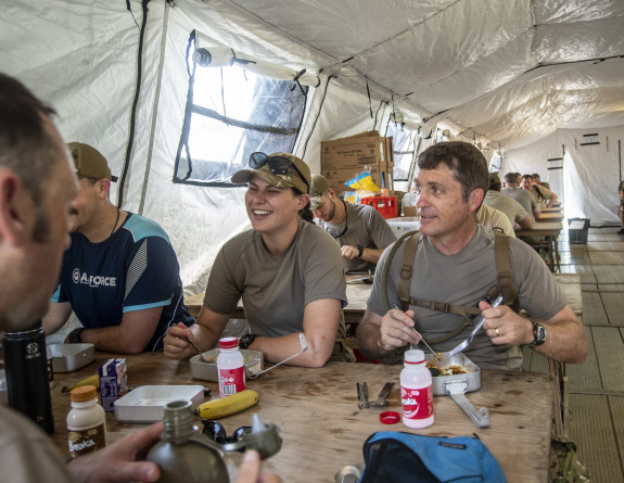 NZDF personnel smile while eating a meal in a large mess tent.