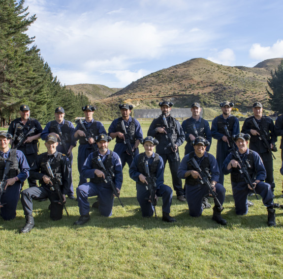 16 sailors in Navy overalls holding weapon stand and take a knee for a group photo on the grass covered range with trees down the sides, and rolling hills in the background.