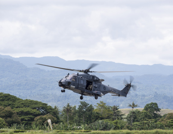 Grey helicopter hovers above the ground, tree and hills are visible in the background.
