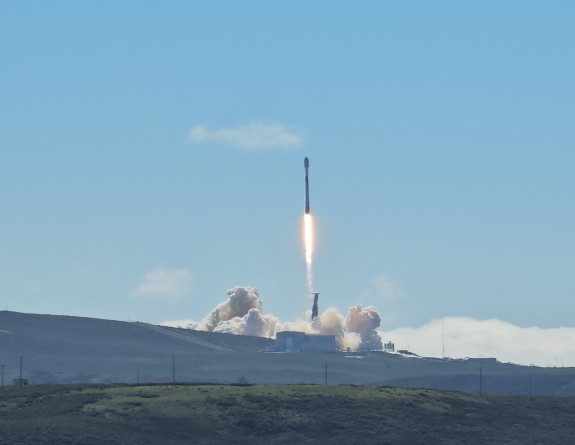 A rocket launching from a hill. In the background is blue sky with some cloud. There is a puff of smoke under the rocket. 
