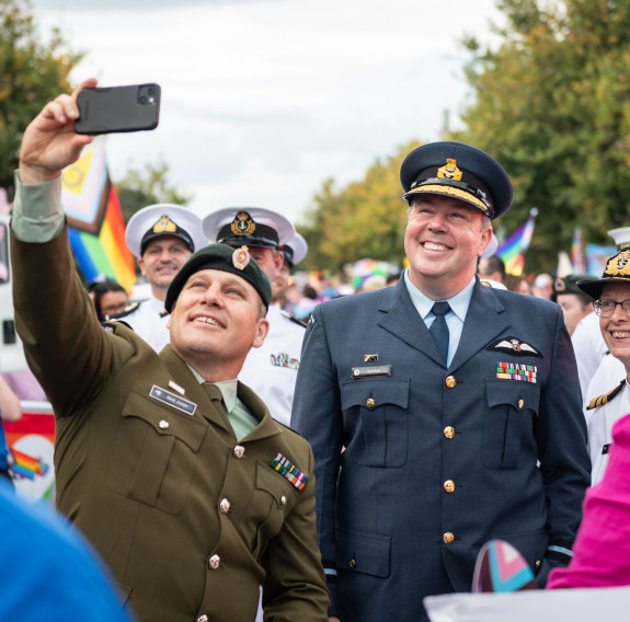 A soldier takes a selfie with an aviator and a sailor at the Auckland Rainbow Parade. In the background is a contingent of sailors followed by pride flags and a tree line to the vanishing point.
