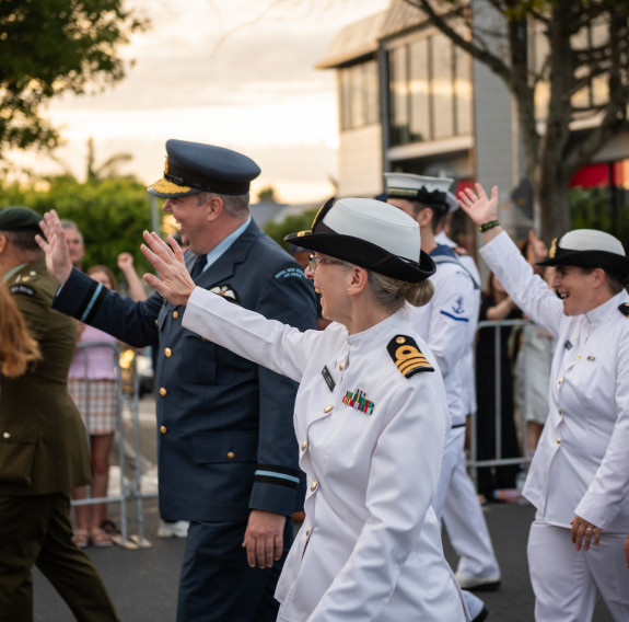 Soldiers, aviators and soldiers wave to the crowd as they pass by. In the background the sun breaks through the clouds and reflects off the windows of a nearby building.