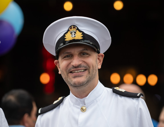 A man in Navy uniform smiles at the camera, small lights are blurred in the background.