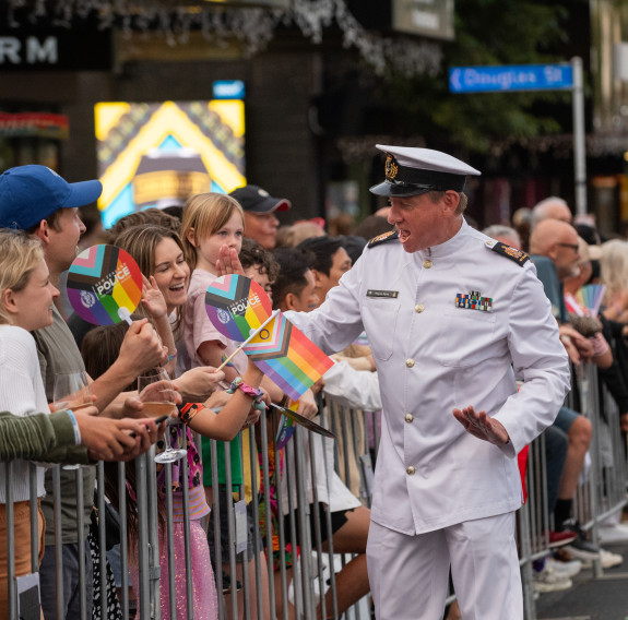 A sailor hi-fives the crowd, behind a metal barricade. They are holding pride flags with looks of joy on their faces.