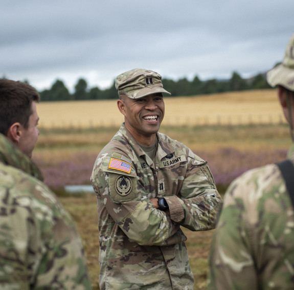 US Army soldier in camo uniform smiles at three other soldiers in the foregound. They stand in front of a field in Waiouru on an overcast day.
