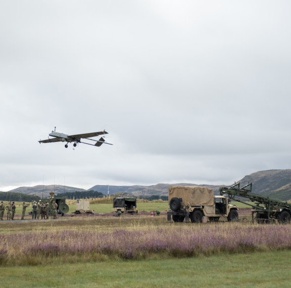 The Shadow UAS just after take off. On the right is the launch ramp and on the left in the background are soldiers standing next to a vehicle with hills in the background. The UAS looks like a small plane, it flies just above the horizon on a cloudy day.