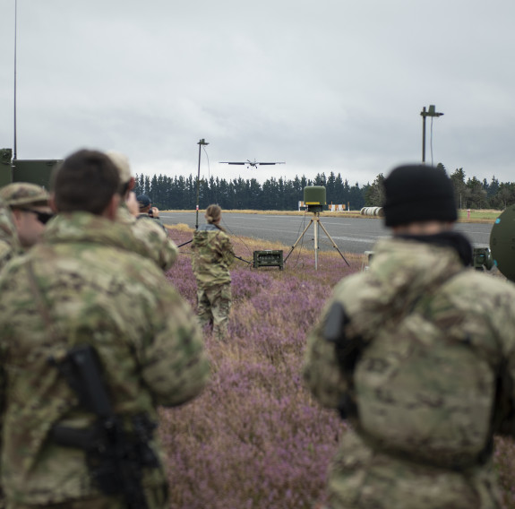 At least five soldiers stand amongst a field of lavender coloured-flowers as the UAV flies down a road, just above the tree line in the background. Lots of towers and monitoring equipment is present around the area.