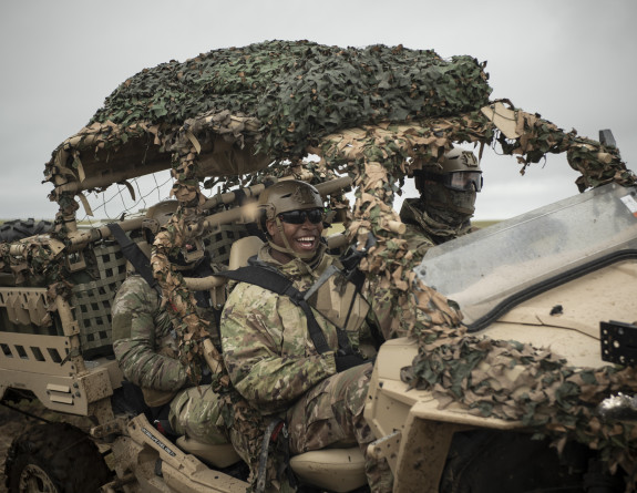 Soldiers smile while driving to site in the MRZR. The MRZR is an all terrain vehicle with a roll cage and is painted tan. Camouflage is wrapped around parts of the vehicle and the soldiers are also wearing camo. 