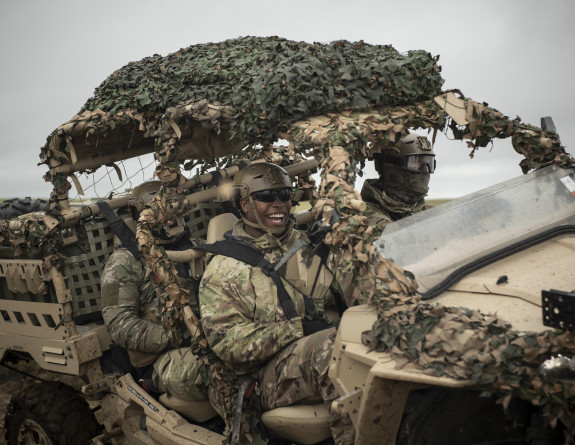 Soldiers smile while driving to site in the MRZR. The MRZR is an all terrain vehicle with a roll cage and is painted tan. Camouflage is wrapped around parts of the vehicle and the soldiers are also wearing camo.