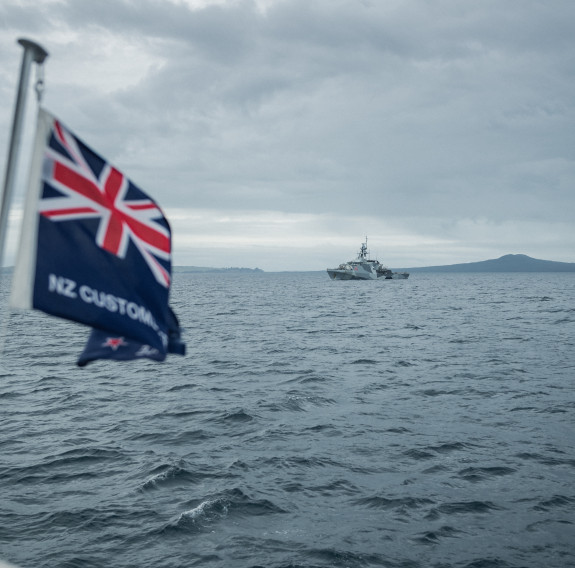 On the left is a New Zealand Customs flag blowing in the wind, a Union Jack is visible. In the background, on the sea in the Hauraki Gulf, HMS Tamar, the Royal Navy ship waits. 