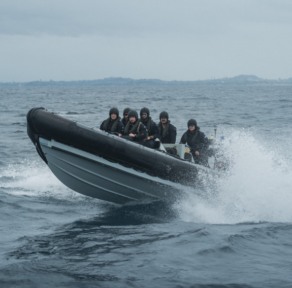 The Deployable Boarding team in a RHIB, preparing to board a vessel of interest during a training exercise. The RHIB is moving fast with water splashing around the stern and the bow out of the water as it moves from right to left.