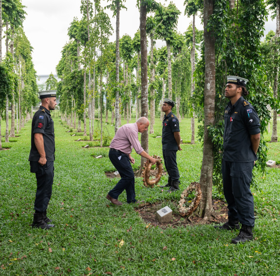 A wreath is placed at the bottom of a tree which stands in neat rows to the horizon. Three Navy sailors stand in a triangle around the wreath laying.