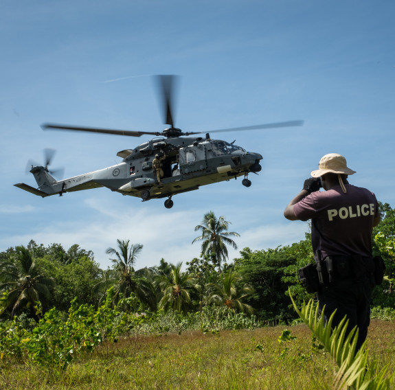 A man wearing a t-shirt and hat with the word "Police" on the back looks as an RNZAF NH90 helicopter comes in to land.