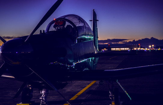 A RNZAF T-6C Texan II training aircraft sits on the tarmac as the sun sets and night begins.
