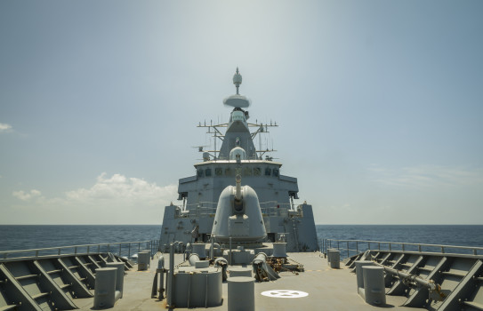 Royal New Zealand Navy frigate, HMNZS Te Kaha is photographed from the bow, showing the large grey 5inch gun and the bridge. On the horizon, some fluffy clouds linger otherwise the conditions are calm and sunn.y 