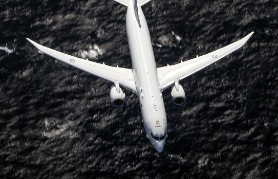 Top down view of a RNZAF P-8A Poseidon, a large grey aircraft with a single jet on each wing. It flies over a dark ocean on exercise.