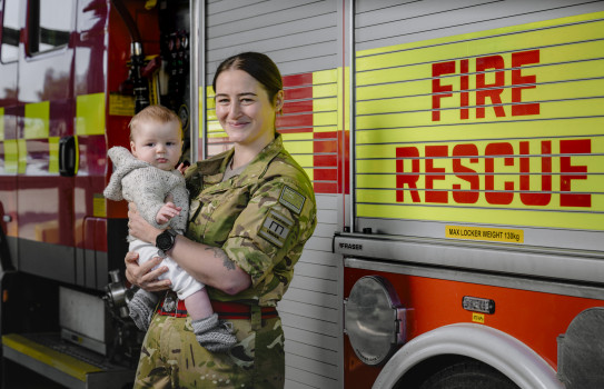 A soldier in uniform holds a baby both are looking at the camera. There's a fire truck in the background which says 'fire rescue' on it. 