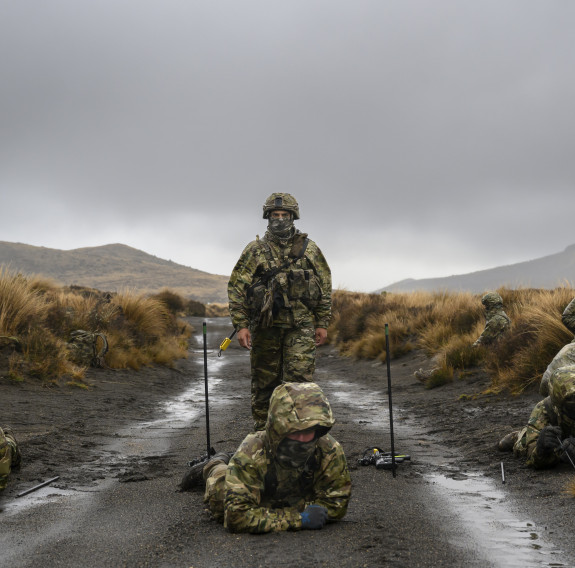 Three soldiers lay belly down on a gravel track as they conduct tactical mine clearance. Each side of the track is surrounded by tussocks under greys skies. An instructors stands behind them watching.